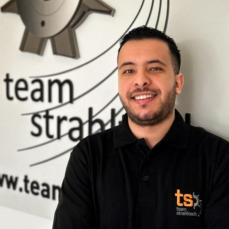 Ein lächelnder Mann in schwarzem Poloshirt vor einer Wand mit "team strathtech"-Logo.