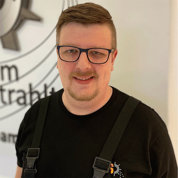 Mann mit Brille, lächelt leicht, trägt schwarzes T-Shirt, Logo im Hintergrund.