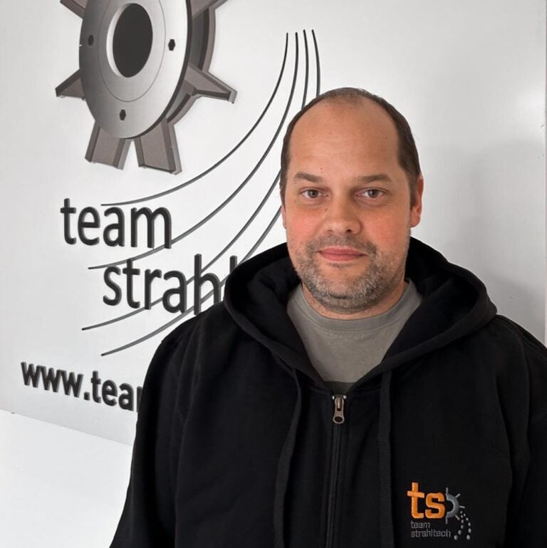 Mann lächelt leicht, trägt schwarzen Hoodie. Im Hintergrund Logo mit Zahnrad, Text "team strahltech".