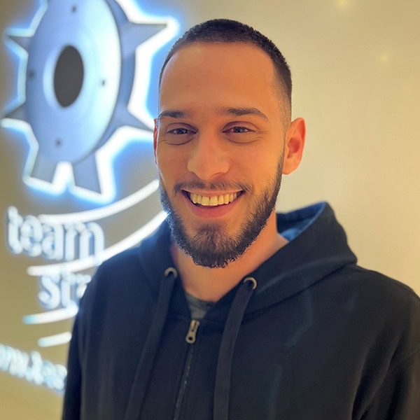 Ein lächelnder Mann mit Bart trägt einen schwarzen Hoodie vor einem beleuchteten Logo.