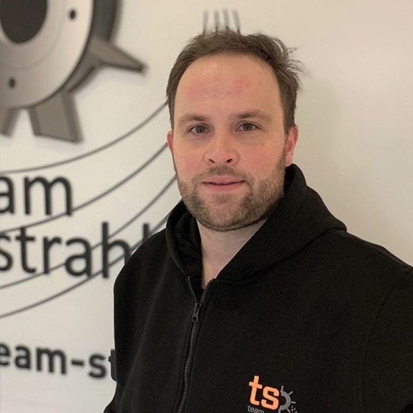 Ein Mann mit einem lenzenden Ausdruck, trägt einen schwarzen Hoodie vor einem technischen Hintergrund.