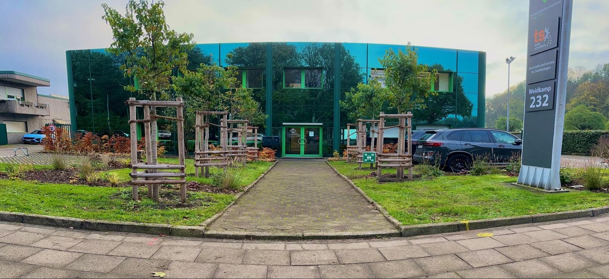 Modernes Gebäude mit grüner Glasfassade, Bäume, gepflegter Garten, geparkte Autos, Firmenschild.