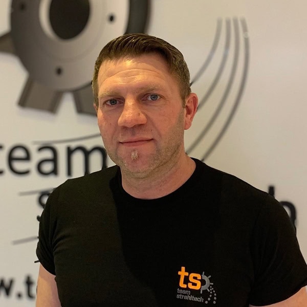 Mann mit neutralem Gesichtsausdruck, schwarzes T-Shirt; Logo im Hintergrund.