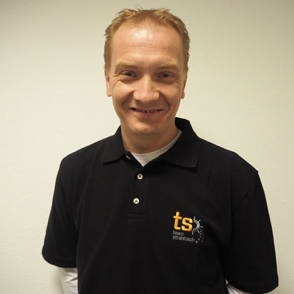 Ein lächelnder Mann in einem schwarzen Polohemd mit gelbem Logo vor einer weißen Wand.