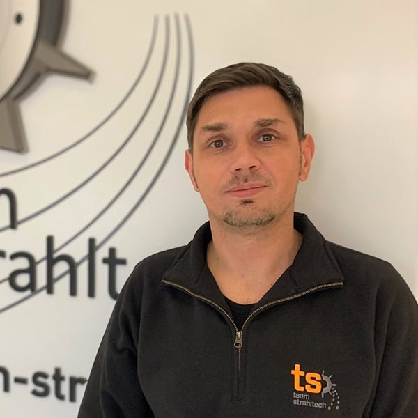 Ein Mann mit ernstem Gesichtsausdruck, trägt schwarzen Pullover mit "ts"-Logo vor weißer Wand.