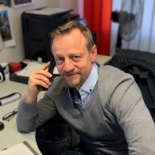 Mann lächelt am Schreibtisch, hält ein Telefon; trägt grauen Pullover über kariertem Hemd.