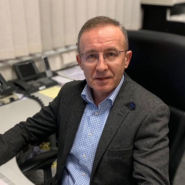 Älterer Mann mit Brille, neutrale Miene, graues Sakko, blaukariertes Hemd, sitzt am Schreibtisch.