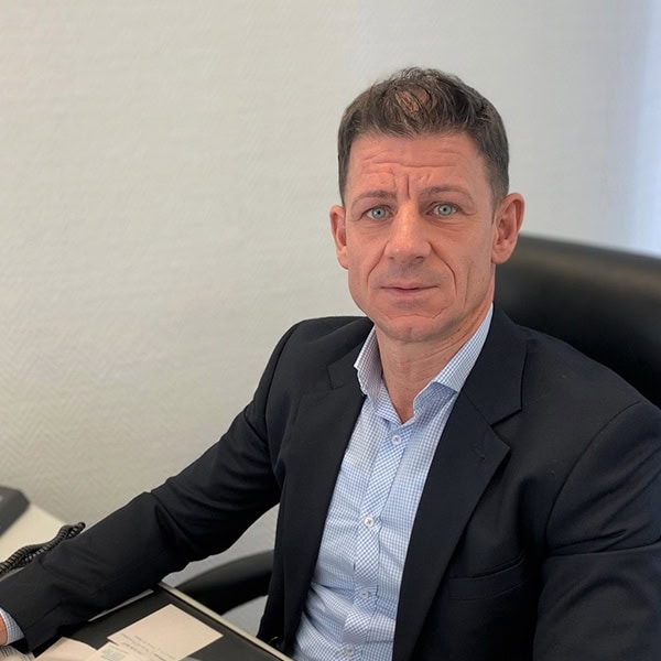 Mann im Anzug sitzt am Schreibtisch, schaut ernst; weißes Hemd, kurze braune Haare, blauer Hintergrund.