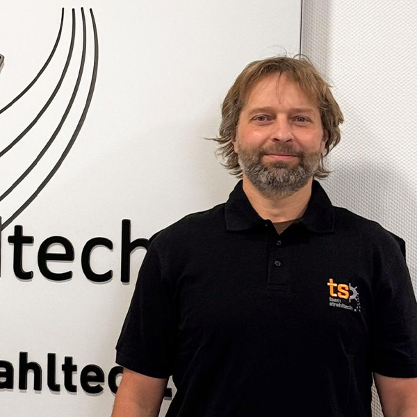 Mann mit Bart lächelt vor einem Schild in schwarzem Polohemd mit "ts" Logo auf der Brust.