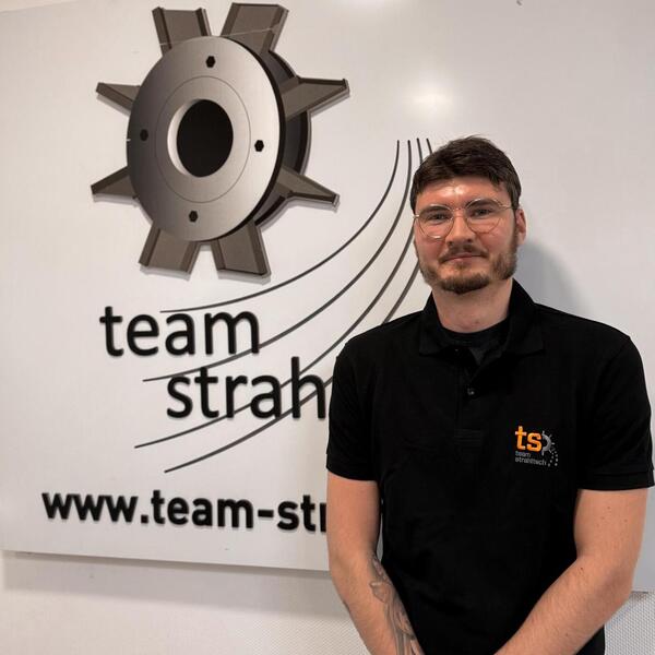 Eine Person mit Brille und Bart lächelt, trägt ein schwarzes Polo mit Team-Logo.