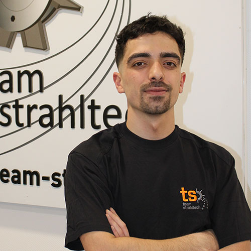 Ein junger Mann mit dunklem Haar und Bart steht lächelnd vor einem weißen Schild