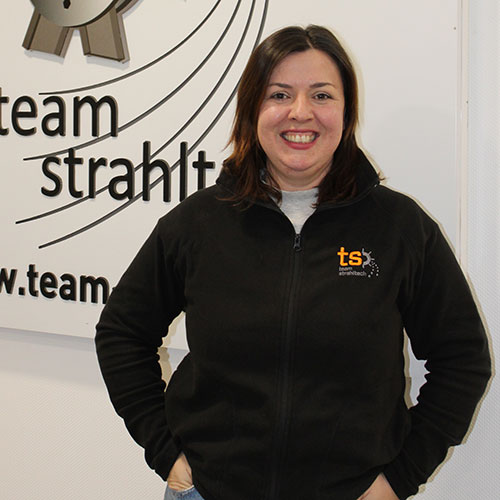 Eine dunkelhaarige Frau lächelt freundlich vor einem weißen Team-Strahlt Schild